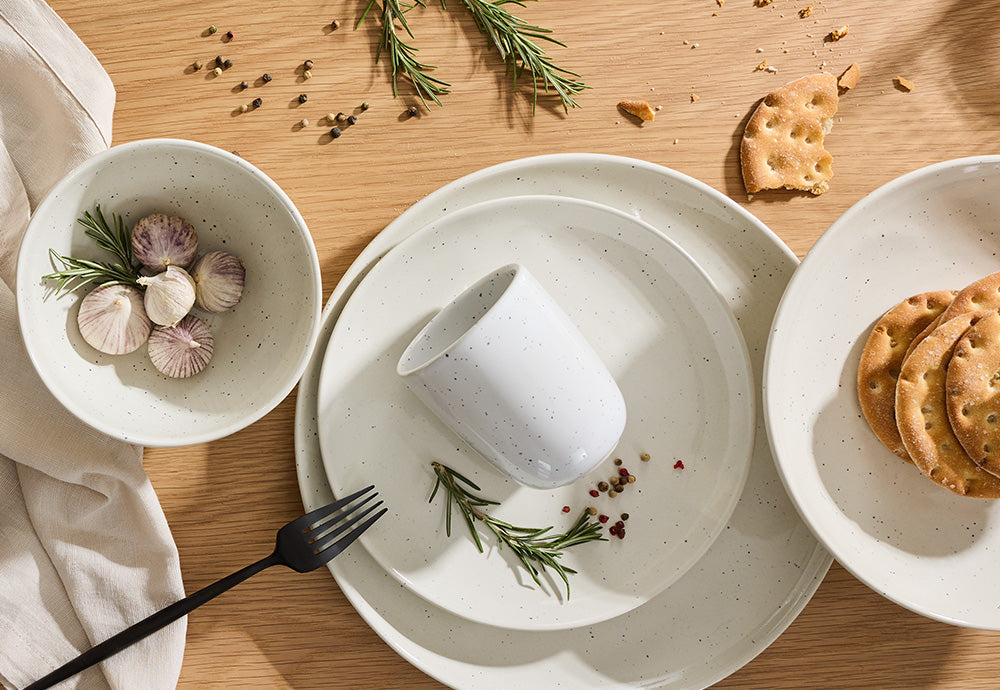 Modernes Geschirr-Set aus weißer Keramik mit feinen schwarzen Sprenkeln, bestehend aus Tellern, Schale und Becher auf einem hellen Holztisch.