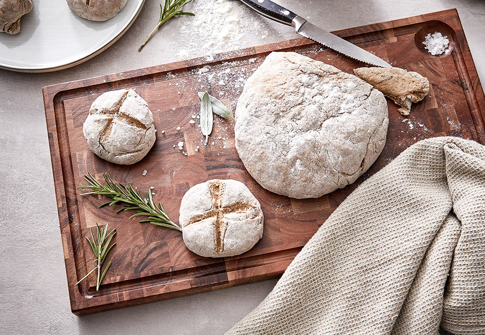 Großes Schneidebrett aus Akazienholz mit Saftrinne, belegt mit frisch gebackenem Brot, Mehl und Rosmarin in einer Landhausküche.