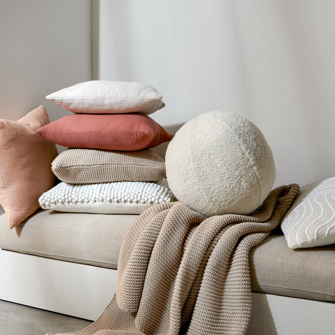 Stapel aus quadratischen Zierkissen in Rosa und Beige sowie ein rundes Bouclé-Kissen auf einer hellen Sitzbank mit Strickdecke.