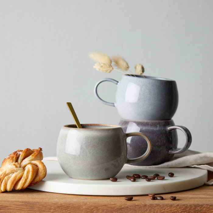 Drei Henkeltassen aus Steinzeug mit blau-grauer Glasur auf einem weißen Serviertablett, dekoriert mit Kaffeebohnen und Gebäck. 