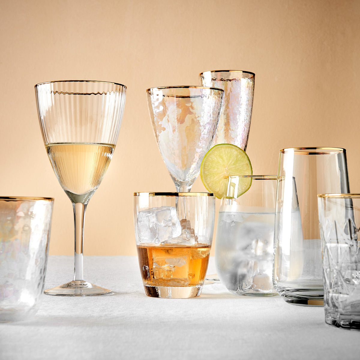 Verschiedene Gläser mit edlem Goldrand – darunter Sekt-, Wasser-, Longdrink- und Weingläser – stilvoll arrangiert auf einer weißen Fläche vor einer cremegelben Wand.